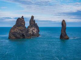 Three unique black rock formations of varying shapes and sizes stand tall in the blue ocean. Off the coast of Iceland under a bright blue sky with white clouds. photo