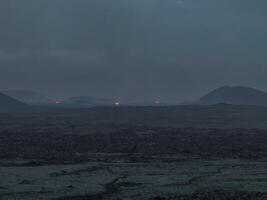 Aerial view of a dark volcanic landscape in Iceland at dusk, featuring small glowing red craters. The sky is a deep blue, contrasting with the black ground. photo
