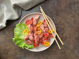 Sauteed sausages with bell peppers served on a plate with green lettuce on a rustic table with chop stick. Top view photo