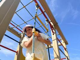 Happy Asian little girl playing rope climbing in playground in summer sunny day. Bottom view photo