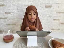 Muslim Asian little girl wearing hijab eating pastry while watching tablet in dining table. Gadget addictive concept. photo