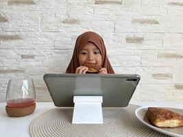 Muslim Asian little girl wearing hijab eating pastry while watching tablet in dining table. Gadget addictive concept. photo