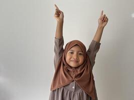 Expressive Asian Muslim little girl wearing hijab looking at the camera and rise two hand with pointing up pose photo