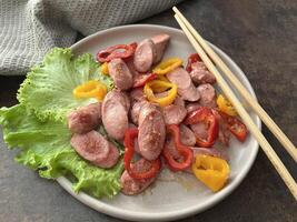 Sauteed sausages with bell peppers served on a plate with green lettuce on a rustic table with chop stick photo