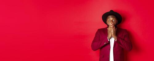 Fashion and party concept. Shocked Black man react on christmas advertisement, gasping and staring at camera with disbelief, standing over red background photo