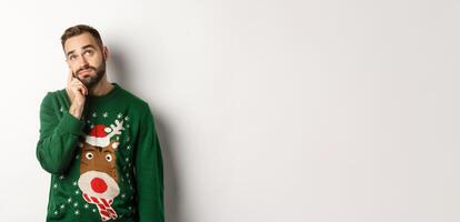 New Year, holidays and celebration. Sad young man with beard, wearing green sweater, showing a teardrop on his face and looking up upset, standing over white background photo
