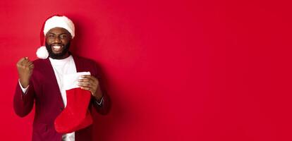 Happy man rejoicing, receive presents in Christmas sock, making fist pump and smiling satisfied, standing over red background photo