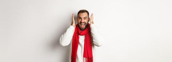 Christmas holidays and New Year concept. Angry man in scream and shout, shaking hands near head with frustrated expression, standing over white background photo