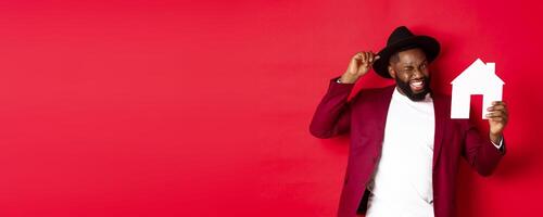 Real estate. Cheerful Black man showing paper house and smiling, recommending broker, standing over red background photo