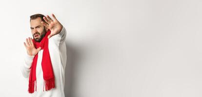 Guy asking to stop, grimacing and pulling away from camera, covering face with hands, standing over white background in red scarf and sweater photo