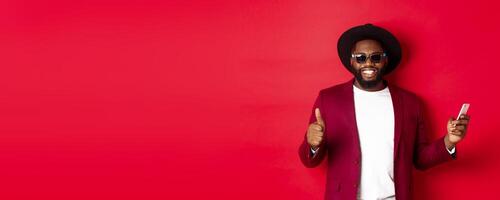Handsome african american man using phone app and showing thumb up, smiling at camera, wearing sunglasses and fancy hat, red background photo