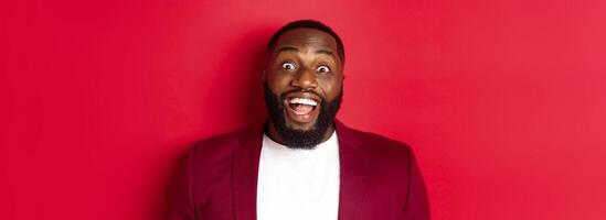 Close-up of african american man with beard staring amazed at camera, smiling and looking excited, standing in blazer against red background photo