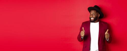 Cheerful Black man having fun on party, showing thumbs up in approval, smiling and liking something, standing against red background photo