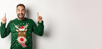 Winter holidays and christmas. Excited man pointing fingers up, showing advertisement, standing over white background photo