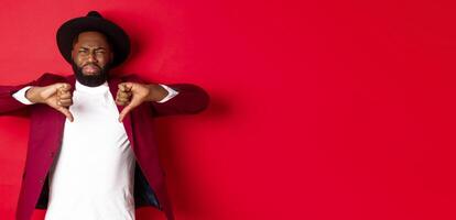 Disappointed Black man leave negative feedback, showing thumb down and grimacing from dislike and disgust, standing in party clothes over red background photo