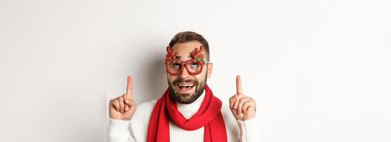Christmas, New Year and celebration concept. Close-up of excited bearded guy in party glasses, pointing and looking up at store promo, white background photo