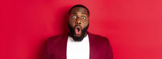 Close-up of shocked Black man gasping and dropping jaw impressed, staring at camera, standing over red background photo