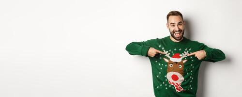 New year celebration and winter holidays concept. Cheerful man pointing at his funny Christmas sweater and smiling, white background photo