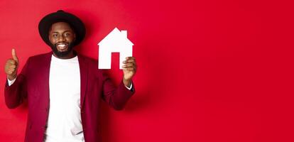 Real estate. Stylish african american bearded man showing home maket, thumb up, recommending agency or apartment complex, standing over red background photo