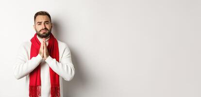Christmas holidays and New Year concept. Desperate man begging for help, asking for favour, holding hands in pray and looking with hope at camera, standing over white background photo