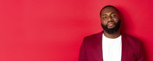Close-up of handsome bearded plump guy smiling at camera shy, standing in stylish blazer on t-shirt, standing over red background photo