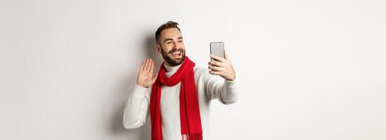 Handsome bearded man calling friends, saying hello and waving hand at mobile phone, standing in sweater and red scarf against white background photo