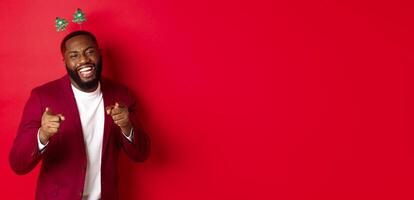 Merry Christmas. Handsome african american man in blazer and party headband, celebrating new year, pointing at camera and smiling, congratulating, red background photo