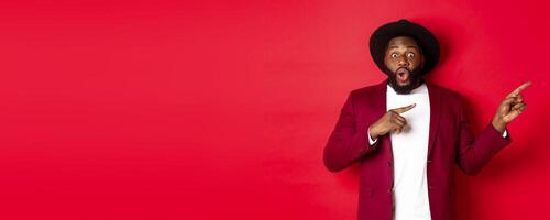 Winter holidays and shopping concept. Stylish african american man pointing fingers right at copy space for logo, standing over red background photo