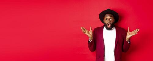 Fashion and party concept. Impressed Black man in classy outfit staring with complete disbelief at camera, gasping and looking surprised, standing over red background photo
