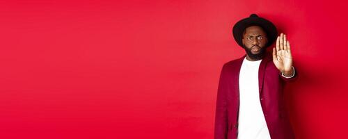 Serious and concerned Black man stretching hand to stop you, prohibit and disagree, frowning and grimacing displeased, standing over red background photo