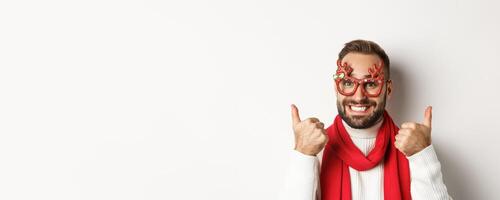 Christmas, New Year and celebration concept. Happy and satisfied man with beard, wearing party glasses, showing thumbs up in approval or like, standing over white background photo