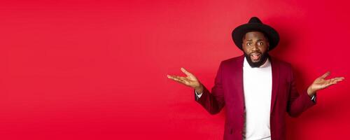 Image of confused Black man asking question, spread hands sideways and staring camera clueless, standing against red background photo
