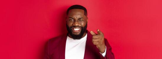 Cheerful Black man with beard wearing party clothes, pointing finger at camera and winking, congratulate with christmas and new year, standing over red background photo
