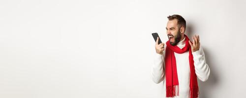 Angry man shouting at smartphone with mad face, standing furious against white background photo