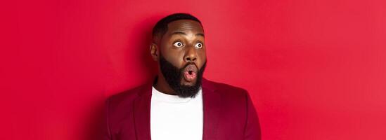 Close-up of impressed Black man looking left at copy space, saying wow and staring with amazement, standing over red background photo