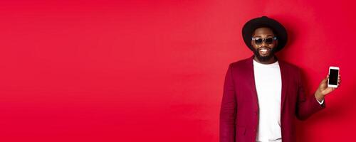 Handsome and stylish Black man showing phone screen at camera, recommending online store or application, standing over red background photo