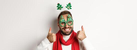Christmas, New Year and celebration concept. Close-up of handsome bearded man in funny party glasses looking happy, showing thumb up in approval, like something, white background photo
