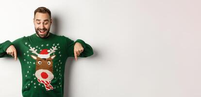 Winter holidays and christmas. Happy bearded man in green sweater, looking and pointing fingers down at advertisement, showing copy space, white background photo