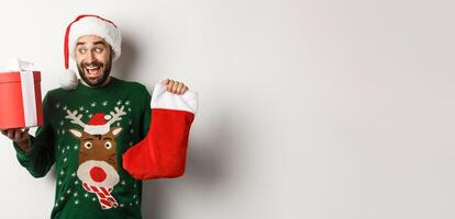 Xmas and winter holidays concept. Happy man got gifts on New Year eve, holding Christmas sock and present inside a box, standing over white background photo