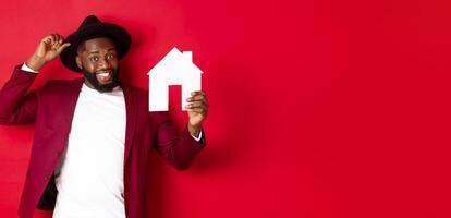 Real estate. Cheerful Black man showing paper house and smiling, recommending broker, standing over red background photo