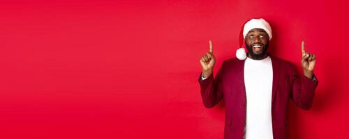Christmas, party and holidays concept. Happy Black man looking at New Year promo, pointing fingers up and smiling, standing over red background photo