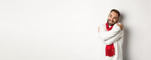 Happy smiling man hugging himself and looking satisfied at upper left corner, wearing warm and comfortable winter sweater, standing over white background photo