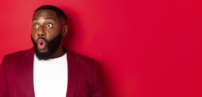 Close-up of impressed Black man looking left at copy space, saying wow and staring with amazement, standing over red background photo