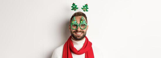 Christmas, New Year and celebration concept. Close-up of handsome bearded man in funny party glasses looking happy, standing against white background photo