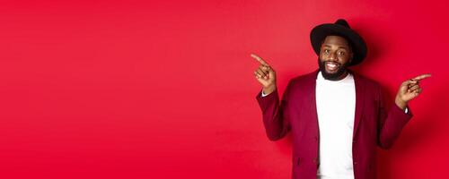 Winter holidays and shopping concept. Cheerful Black man smiling and showing two promo, pointing fingers sideways at copy spaces, red background photo