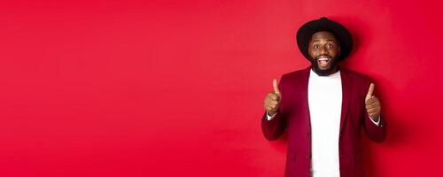 Christmas shopping and people concept. Handsome Black man smiling satisfied, showing thumbs-up, like and agree, approve something, standing against red background photo