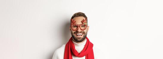 Christmas, New Year and celebration concept. Close-up of happy bearded man smiling, wearing party glasses and red scarf, white background photo