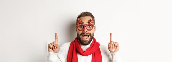 Christmas, New Year and celebration concept. Close-up of handsome bearded man in party glasses and red scarf, celebrate winter holidays and pointing fingers up at copy space photo