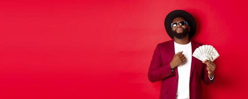 Sassy and cool african american man in sunglasses and hat, pointing at himself and showing dollars, looking confident against red background photo