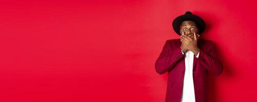 Christmas shopping and people concept. Shocked black man looking startled at camera, covering mouth with hands, standing against red background photo